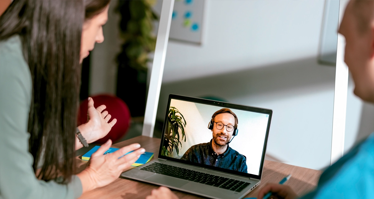 Bei einem hybriden Termin sprechen zwei Personen im Büro mit einem per Video zugeschalteten Kollegen